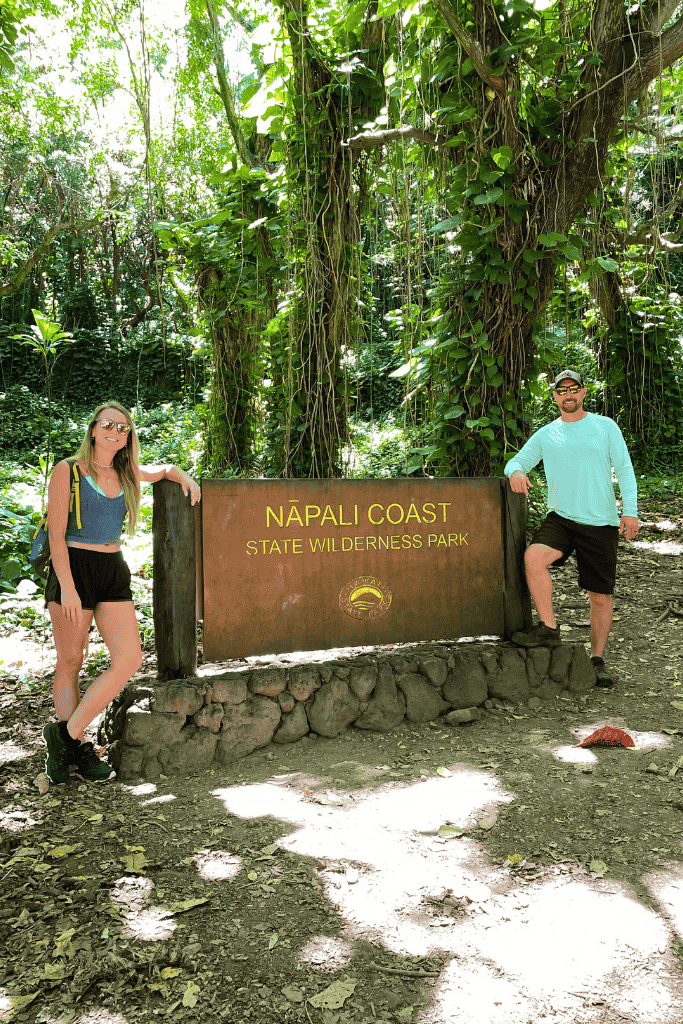 Kate and her companion pose on either side of the Nā Pali Coast State Wilderness Park sign, surrounded by lush jungle foliage and dappled sunlight.