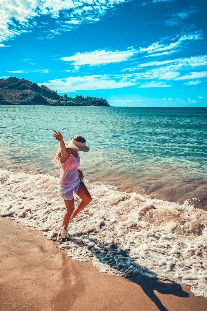 Kate dances playfully in the foamy shoreline wearing a sunhat and cover-up, with turquoise waters and lush Kauai cliffs in the distance.
