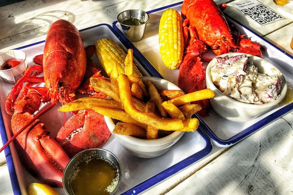 Two trays of classic Maine lobster dinners, featuring whole steamed lobsters, buttered corn on the cob, crispy fries, and creamy potato salad, with melted butter on the side for dipping. The vibrant red lobsters and golden fries are served on simple white trays, capturing the essence of a fresh seafood meal, often enjoyed during fall vacations along the coast of Maine.