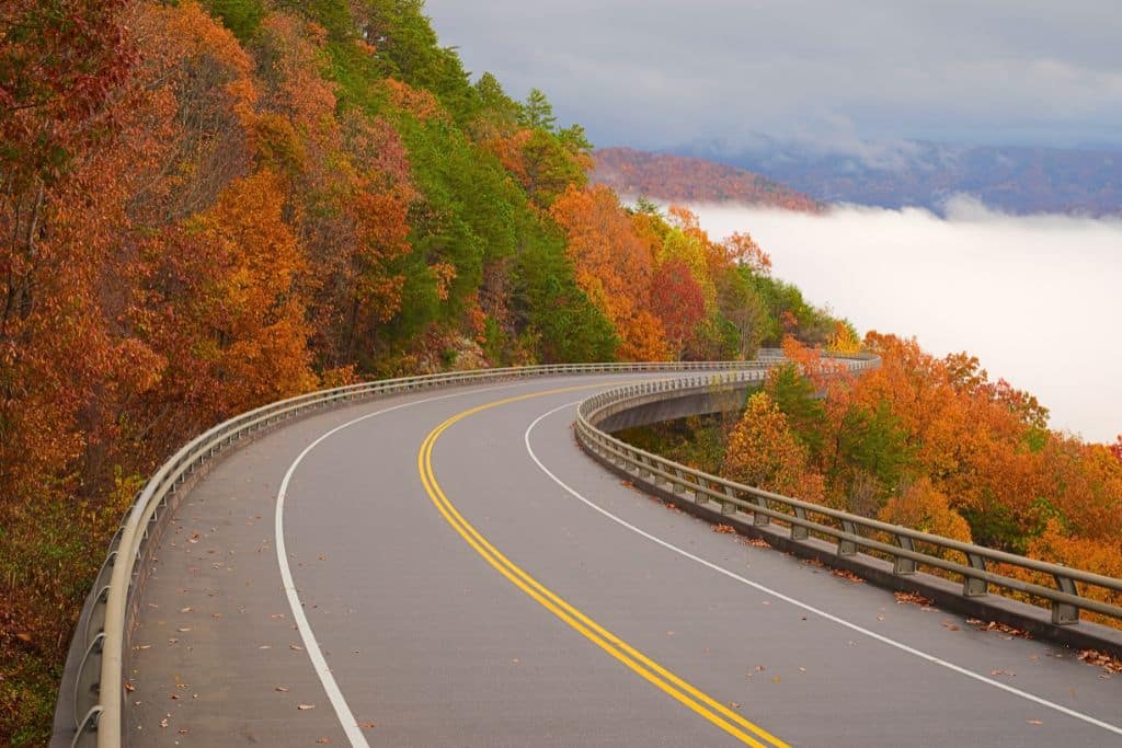 A curving mountain road in the Smoky Mountains, lined with trees in full autumn colors of orange, red, and green. The road winds along a ridge, with clouds or mist blanketing the valleys below, creating a serene and picturesque scene.