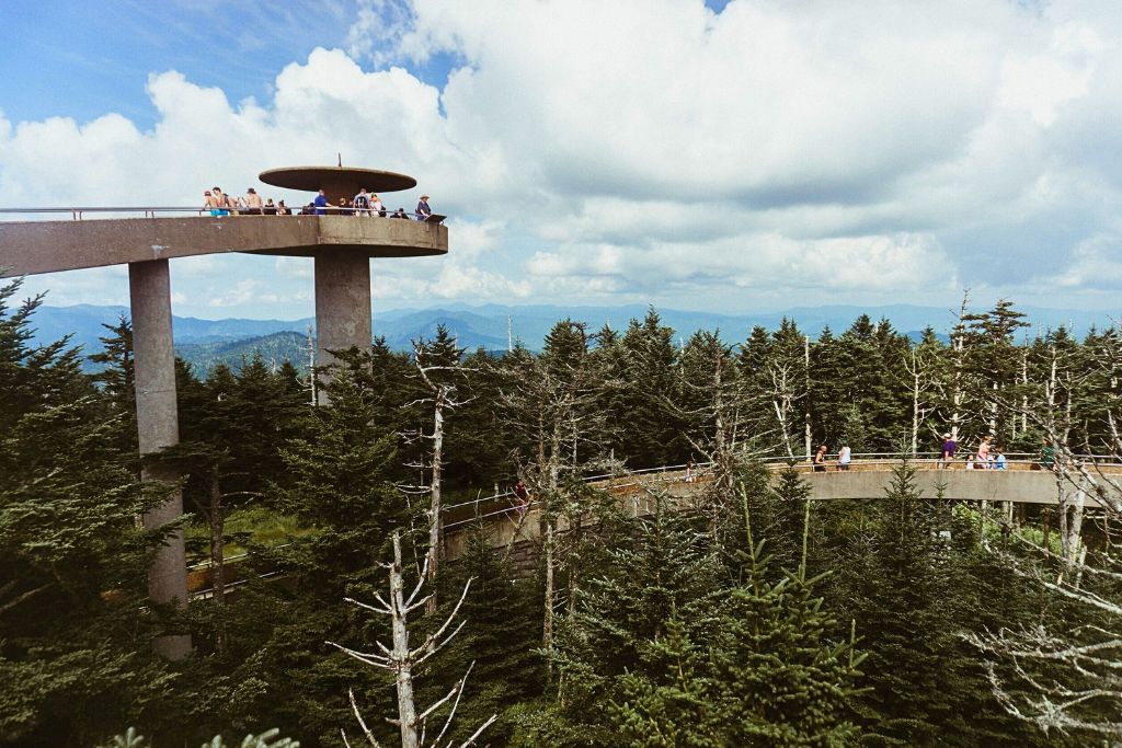 Visitors stand on the circular observation tower at Clingmans Dome, the highest point in the Smoky Mountains, offering panoramic views of the surrounding forest and distant mountain ranges. A curved walkway leads through evergreen trees to the top, under a partly cloudy sky.