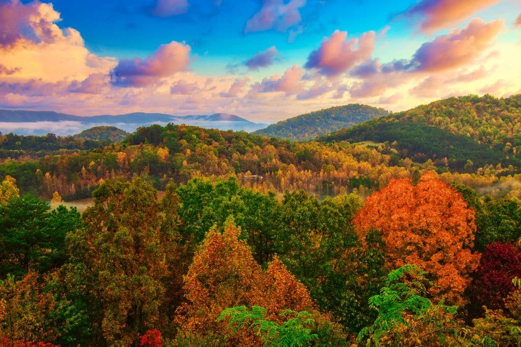 A breathtaking view of rolling hills in the Smoky Mountains covered in vibrant autumn foliage, with shades of green, orange, and red. The sky above is painted with dramatic clouds illuminated by the setting sun, casting a golden glow over the landscape.