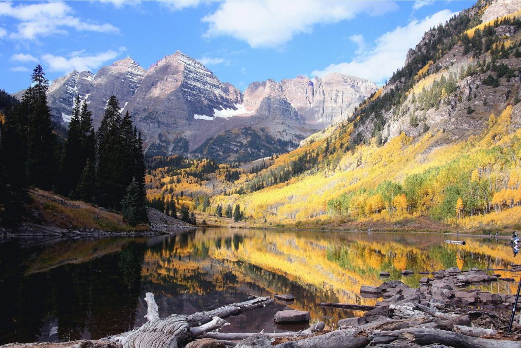 A serene alpine lake reflects the golden yellow aspens and evergreen trees on the surrounding mountainsides, with majestic snow-capped peaks towering in the background. Logs and rocks line the shoreline, enhancing the peaceful natural beauty of the scene.