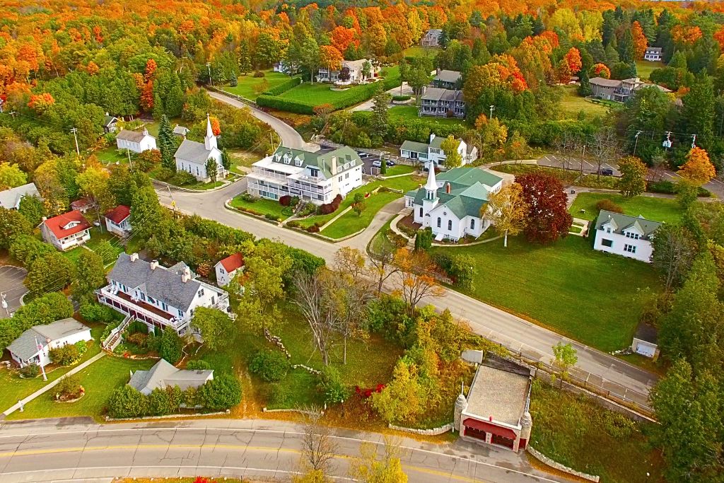 An aerial view of a small town in Door County, Wisconsin, surrounded by vibrant fall foliage. The quaint town features charming houses, a church with a white steeple, and tree-lined streets, all set against a backdrop of colorful autumn trees in shades of orange, yellow, and red. This scene captures the peaceful beauty of fall vacations in Door County, known for its picturesque small towns and stunning seasonal colors.