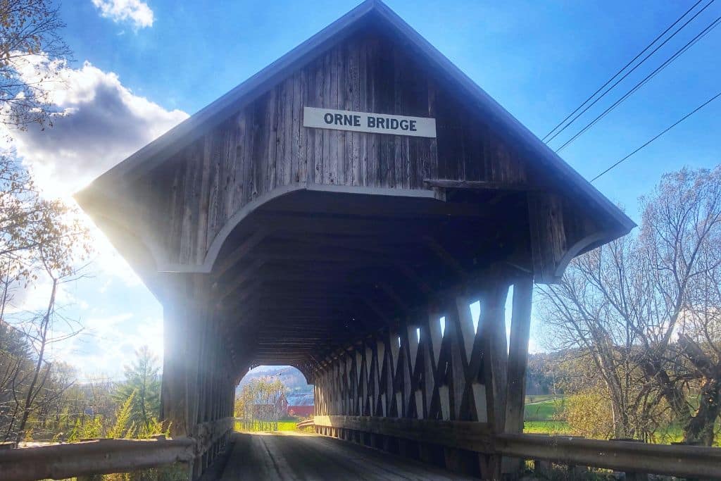 A rustic wooden covered bridge labeled "Orne Bridge" set against a bright blue sky with wisps of clouds. Sunlight filters through the trees, illuminating the interior of the bridge and the scenic green and autumn-colored landscape beyond.