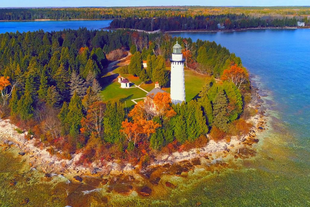 An aerial view of a lighthouse on the edge of a small wooded peninsula surrounded by clear blue water. The trees, displaying autumn colors of orange and yellow, line the shoreline and contrast with the evergreen trees.