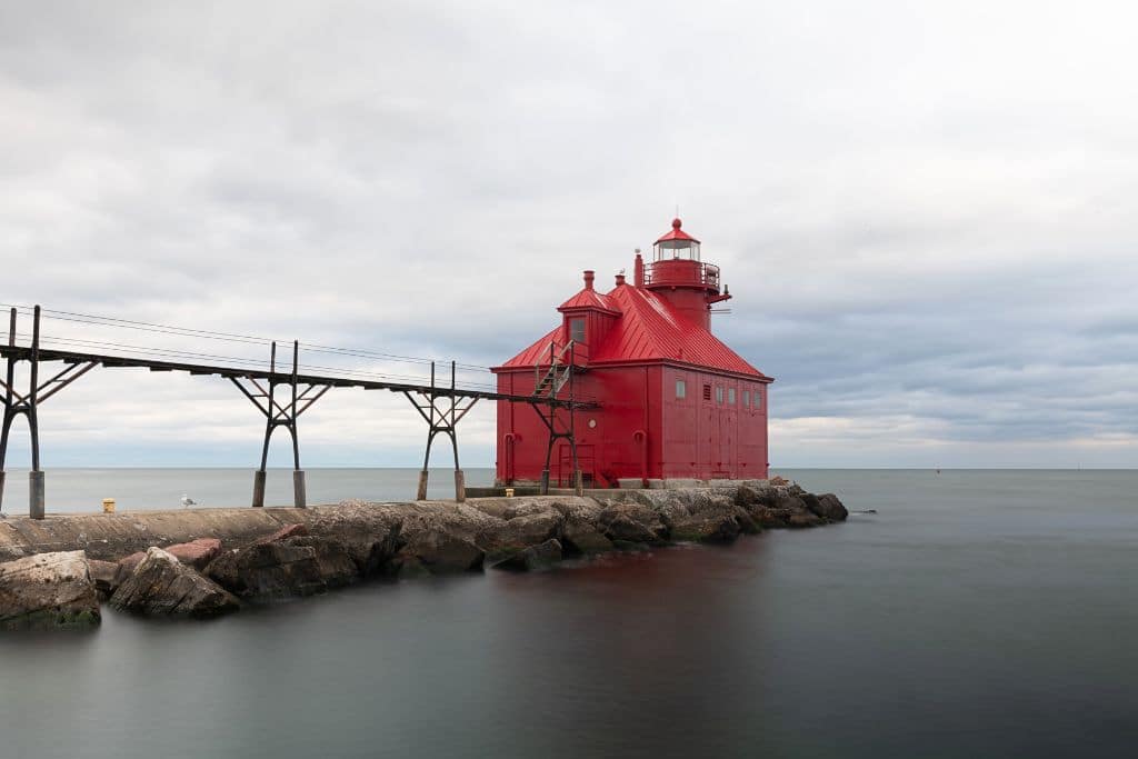 A striking red lighthouse sits at the end of a rocky pier in Door County, Wisconsin, extending into calm waters under a cloudy sky. The lighthouse is connected to the mainland by a metal walkway, adding to its industrial yet picturesque charm.