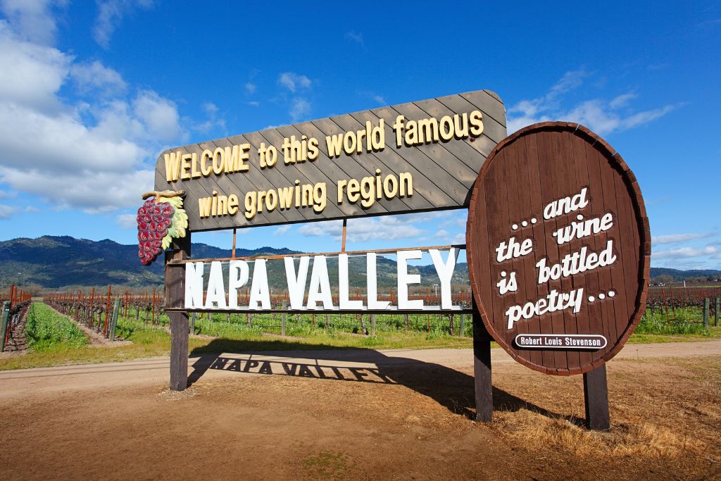 A large wooden sign welcomes visitors to Napa Valley, the world-famous wine-growing region, with the message "...and the wine is bottled poetry," a quote by Robert Louis Stevenson. The sign is set against a backdrop of vineyard rows stretching into the distance, with mountains and a bright blue sky in the background.