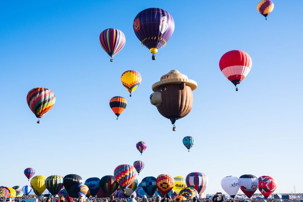 A sky filled with colorful hot air balloons of various shapes and designs during the Albuquerque International Balloon Fiesta. One prominent balloon is shaped like Smokey the Bear, while others display vibrant patterns and colors. Below, crowds gather to watch the mass ascension under a clear blue sky, capturing the excitement of this iconic fall event in Albuquerque, New Mexico.