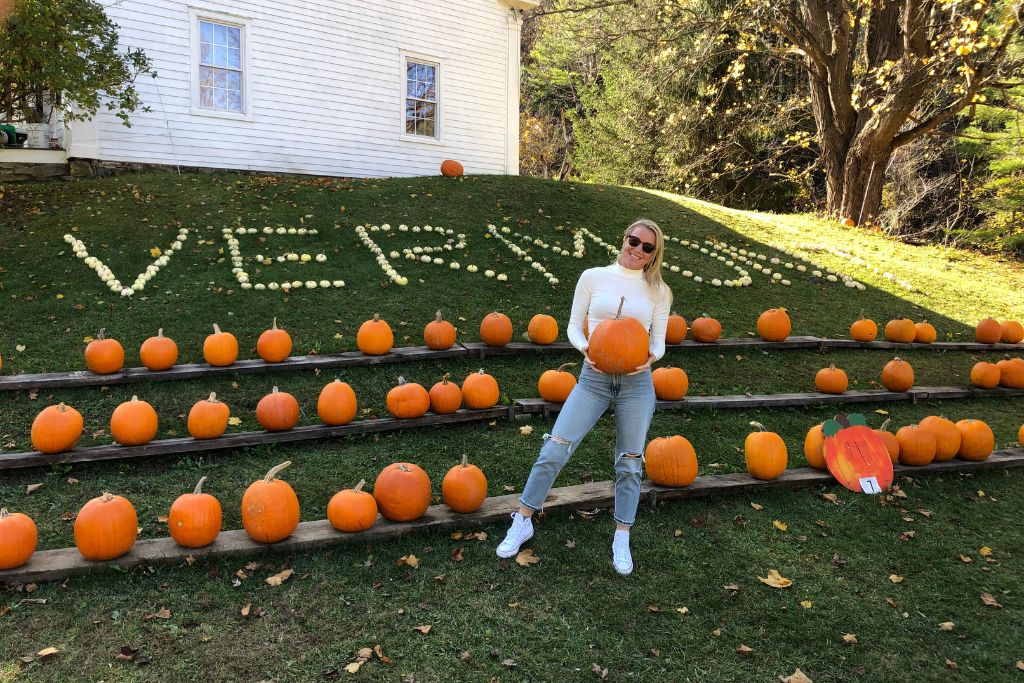 Kate from Kate's Crossing Blog is wearing sunglasses and a white sweater poses with a pumpkin in front of a display of neatly arranged pumpkins on wooden stands. Behind her, white gourds spell out "Vermont" on a grassy hillside, with a white house and trees completing the autumn scene.