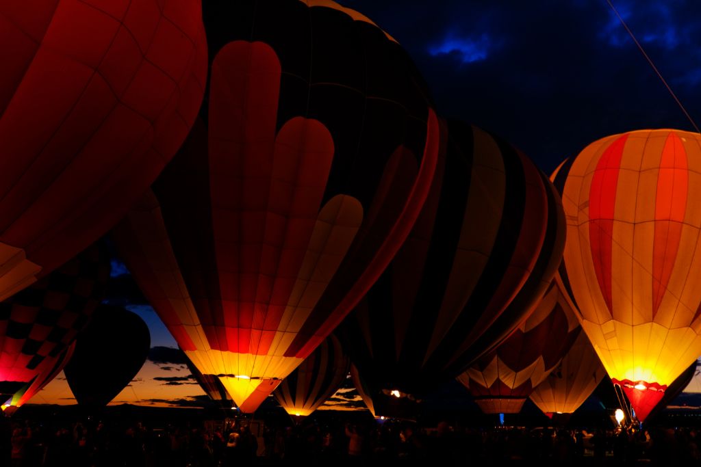 Hot air balloons glow in the dark during the evening at the Albuquerque International Balloon Fiesta. The illuminated balloons create a striking contrast against the dark sky, with their vibrant colors glowing warmly from within. This scene captures the magical atmosphere of the Balloon Glow event.