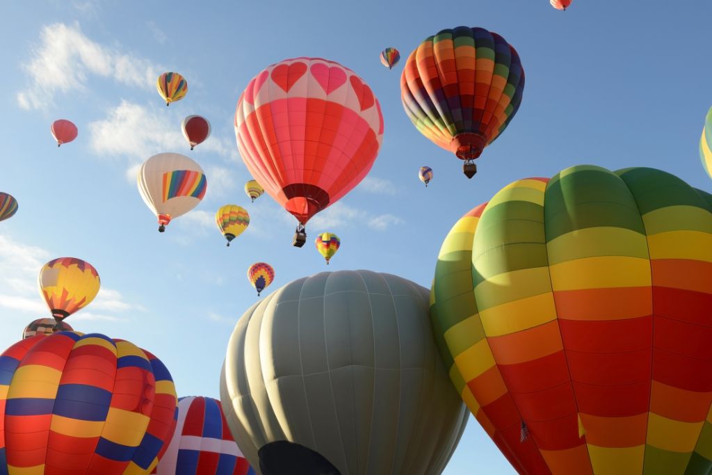 A colorful array of hot air balloons rise into the sky during the Albuquerque International Balloon Fiesta. Balloons of various patterns and designs, including one with red heart shapes, float gracefully under a bright blue sky with scattered clouds.