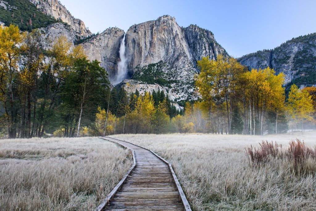 A scenic boardwalk leads through a frost-covered meadow in Yosemite National Park, framed by autumn trees with golden leaves. In the distance, towering granite cliffs loom, with a waterfall cascading down the center. The crisp morning air is highlighted by light mist rising from the ground, adding to the serene, early-morning atmosphere.