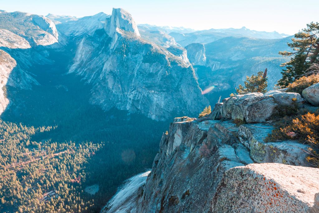 A breathtaking view of Yosemite's Half Dome, rising prominently from the valley floor, framed by rugged granite cliffs and dense forest below. The scene captures the expansive beauty of the Sierra Nevada mountains under clear skies, with the sun casting soft light on the rock formations and valley below.