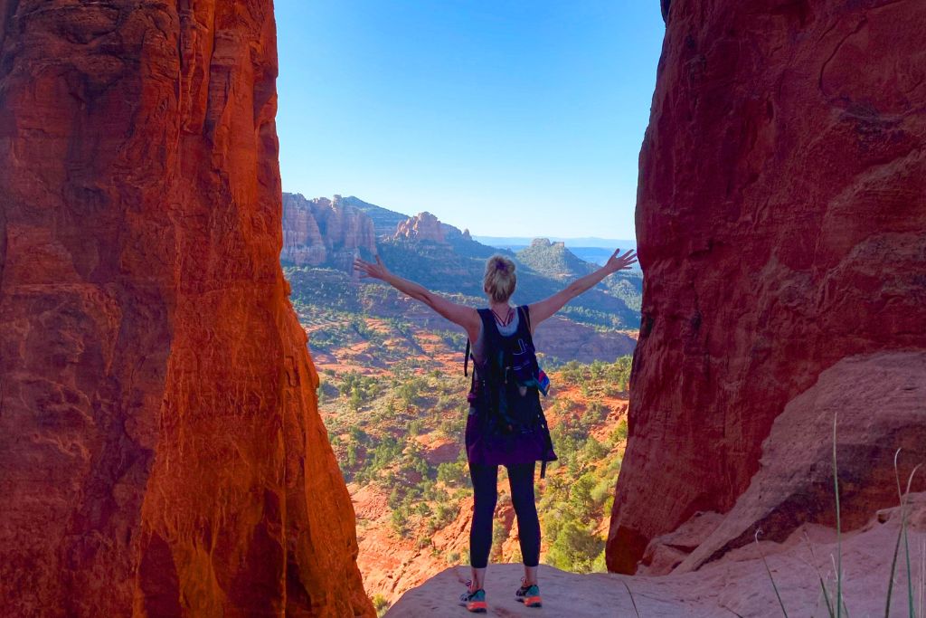 Kate from Kate's Crossing Blog stands with arms outstretched between two towering red rock formations in Sedona, Arizona, looking out at the stunning view of red cliffs and greenery in the distance under a clear blue sky. The vibrant red rocks frame the expansive desert landscape, creating a sense of awe and freedom.