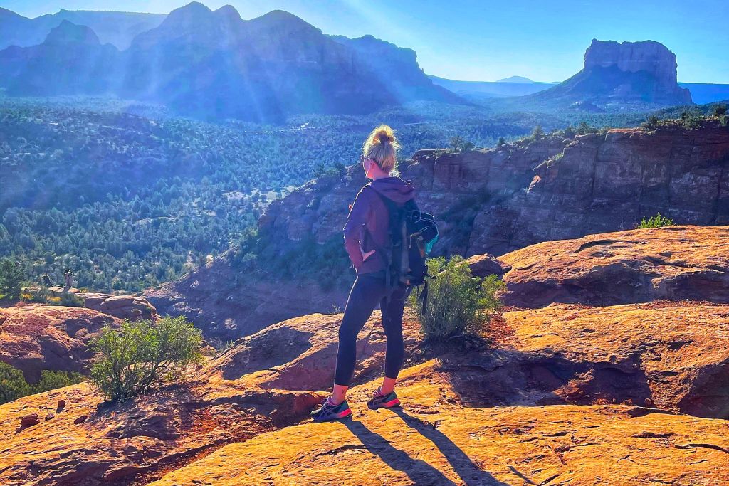 Kate from Kate's Crossing Blog with a backpack stands on a sunlit rocky ledge in Sedona, Arizona, gazing at the expansive desert landscape filled with red rocks, green shrubbery, and distant mountains under a clear blue sky. The sunlight casts soft rays, adding warmth and depth to the scenic view.