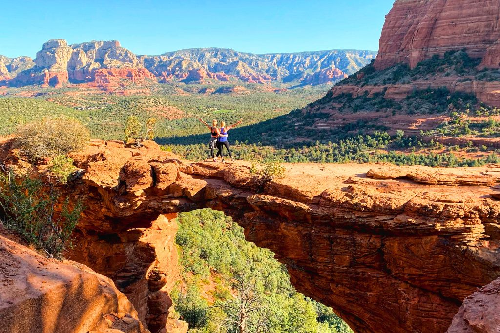 Two hikers stand triumphantly on Devil's Bridge, a natural rock formation in Sedona, Arizona, with stunning red rock cliffs and a green valley sprawling into the distance. The bright blue sky contrasts with the vibrant red hues of the desert landscape, making for a breathtaking scene of adventure and natural beauty.