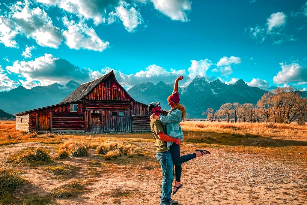 Kate and her husband pose joyfully in front of a rustic wooden barn at Grand Teton National Park, with the man lifting the woman in the air. Behind them, the rugged Teton mountain range rises under a bright blue sky filled with scattered clouds, while the golden autumn grass surrounds them, adding warmth to the picturesque scene.