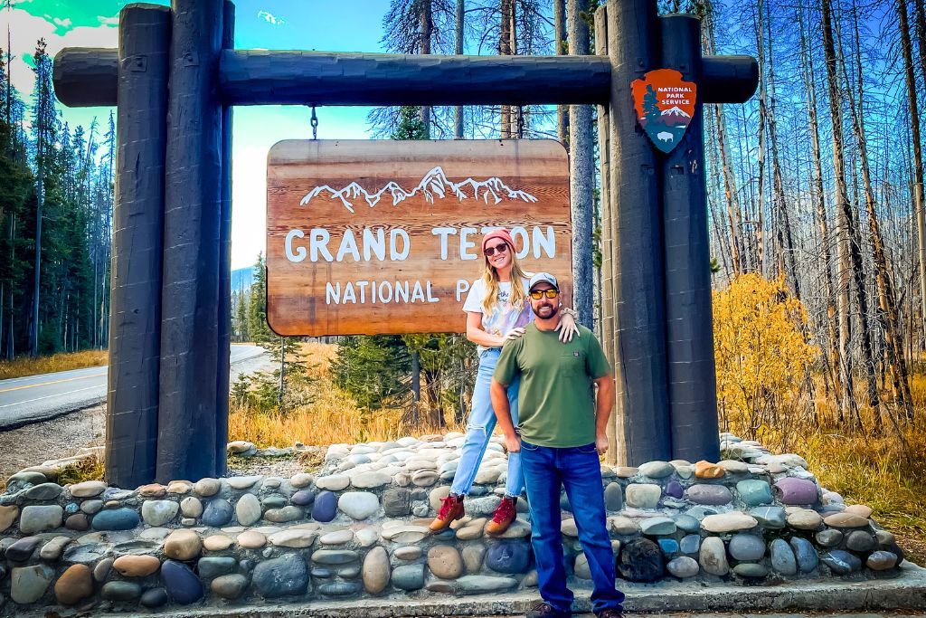 Kate and her husband pose in front of the Grand Teton National Park entrance sign. The wooden sign displays the park's name and a graphic of mountain peaks, framed by tall timber beams and a National Park Service emblem. The surrounding autumn landscape features a road lined with trees, with some foliage turning golden.