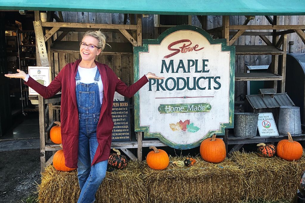 Kate from Kate's Crossing Blog smiles and gestures while standing in front of a large sign that reads "Stowe Maple Products, Home Made." She is dressed casually in overalls and a red cardigan, with pumpkins and hay bales decorating the area.