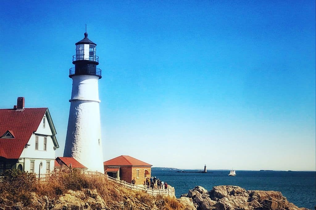 A picturesque lighthouse stands tall on a rocky coastline, overlooking a calm blue ocean under a clear sky. A small group of visitors walks along the path beside the lighthouse, and in the distance, a sailboat glides on the water. This coastal scene, with its iconic New England lighthouse, reflects the tranquil beauty of fall vacations in the US, particularly along the coast of Maine.