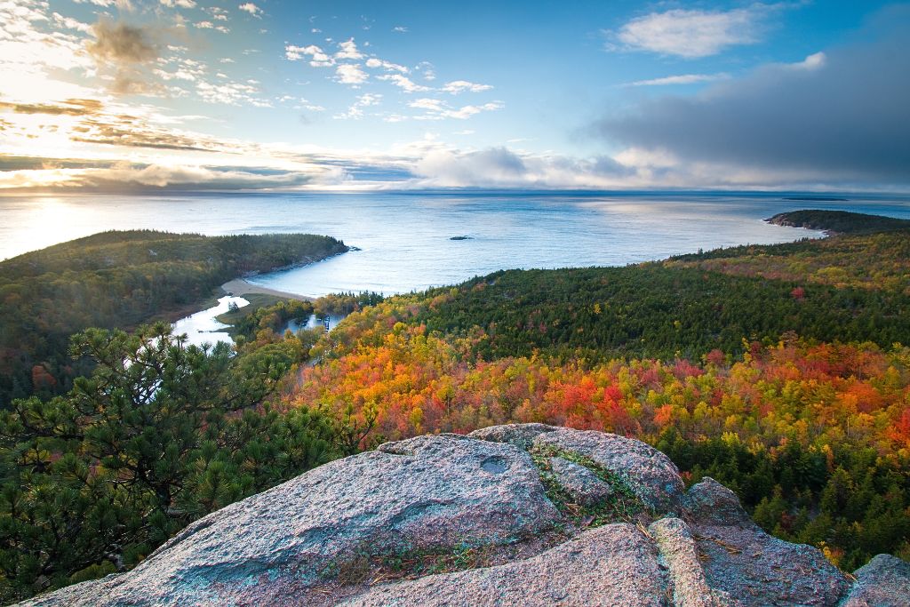 A stunning view from a rocky cliff overlooking Acadia National Park during fall, with colorful autumn foliage covering the forest below. The landscape stretches out to meet the calm waters of the Atlantic Ocean, with a small inlet visible along the coastline. The mix of vibrant trees and serene ocean highlights the natural beauty of Acadia, a popular destination for fall vacations in the US.