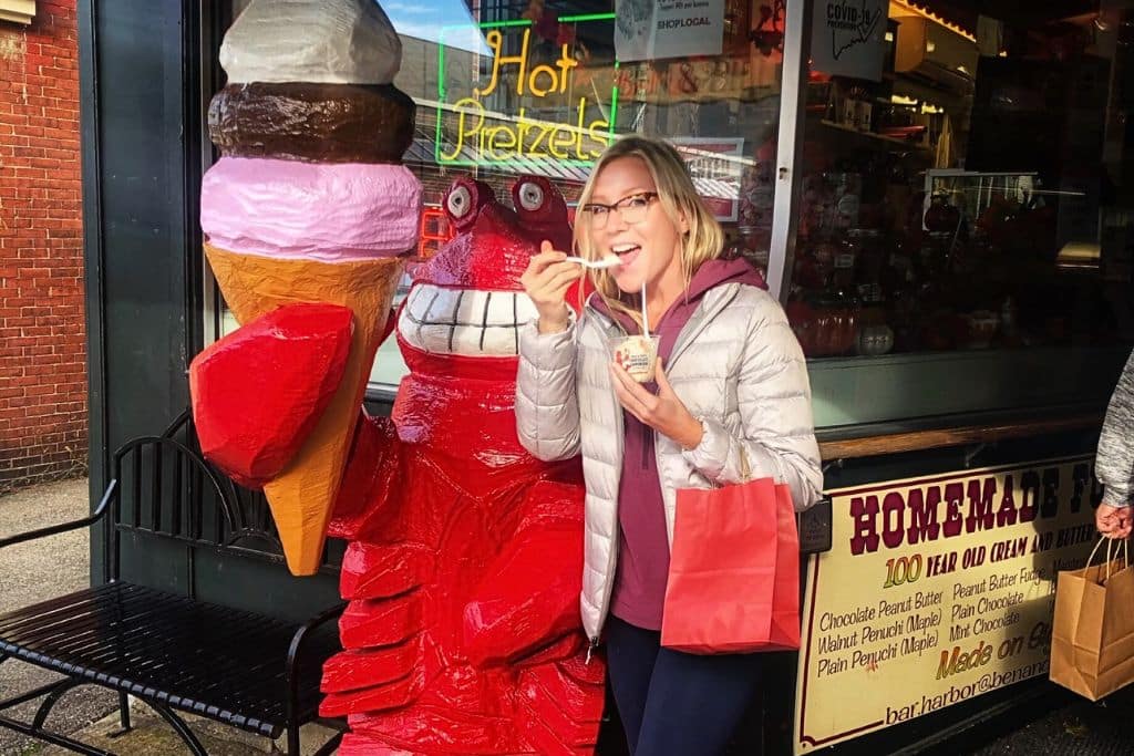 Kate from Kate's Crossing blog, bundled in a jacket, smiles while trying lobster ice cream outside a shop in Bar Harbor, Maine. She stands next to a large red lobster statue holding an ice cream cone, with neon signs and a display advertising homemade fudge in the background.