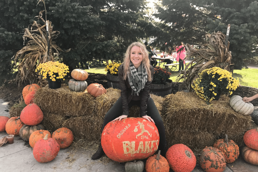 Kate from Kate's Crossing Blog is smiling and sitting on a large orange pumpkin surrounded by a display of various pumpkins and yellow chrysanthemums. The scene is set outdoors with hay bales, corn stalks, and evergreen trees in the background, creating a festive, autumnal atmosphere. This image evokes the spirit of fall vacations in the US, especially at a pumpkin patch or orchard.