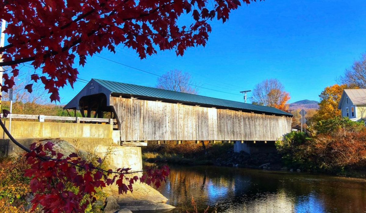 The Cornish-Windsor Covered Bridge over a calm river, framed by vibrant red autumn leaves and surrounded by the scenic beauty of New England in the fall.