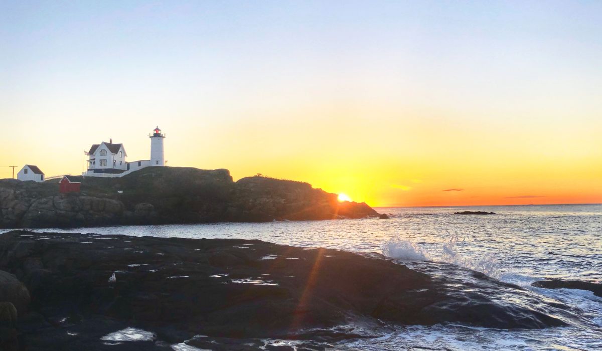 The Nubble Lighthouse in York, Maine, captured at sunrise. The lighthouse stands on a rocky island with waves gently crashing against the shore, and the sky is painted with warm hues of orange and yellow as the sun rises on the horizon.