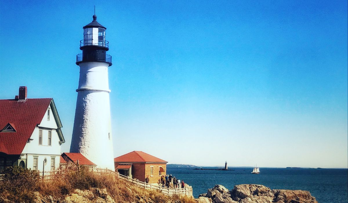 The Portland Head Lighthouse in Maine, standing tall against a clear blue sky. The historic white lighthouse with a black top is adjacent to a red-roofed keeper's house, overlooking the rocky coastline and the expansive ocean. A sailboat is visible in the distance.