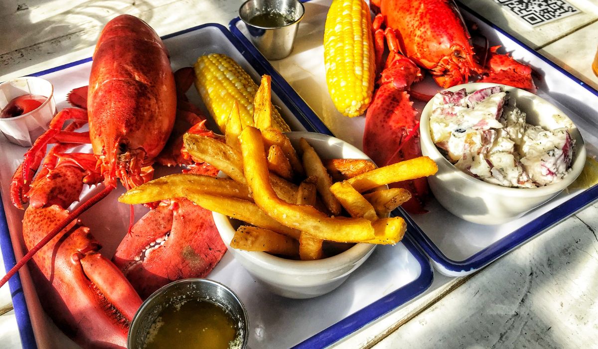 A lobster lunch in Portland, Maine, featuring two trays with whole lobsters, corn on the cob, French fries, and a bowl of potato salad. The trays also include melted butter and dipping sauces.