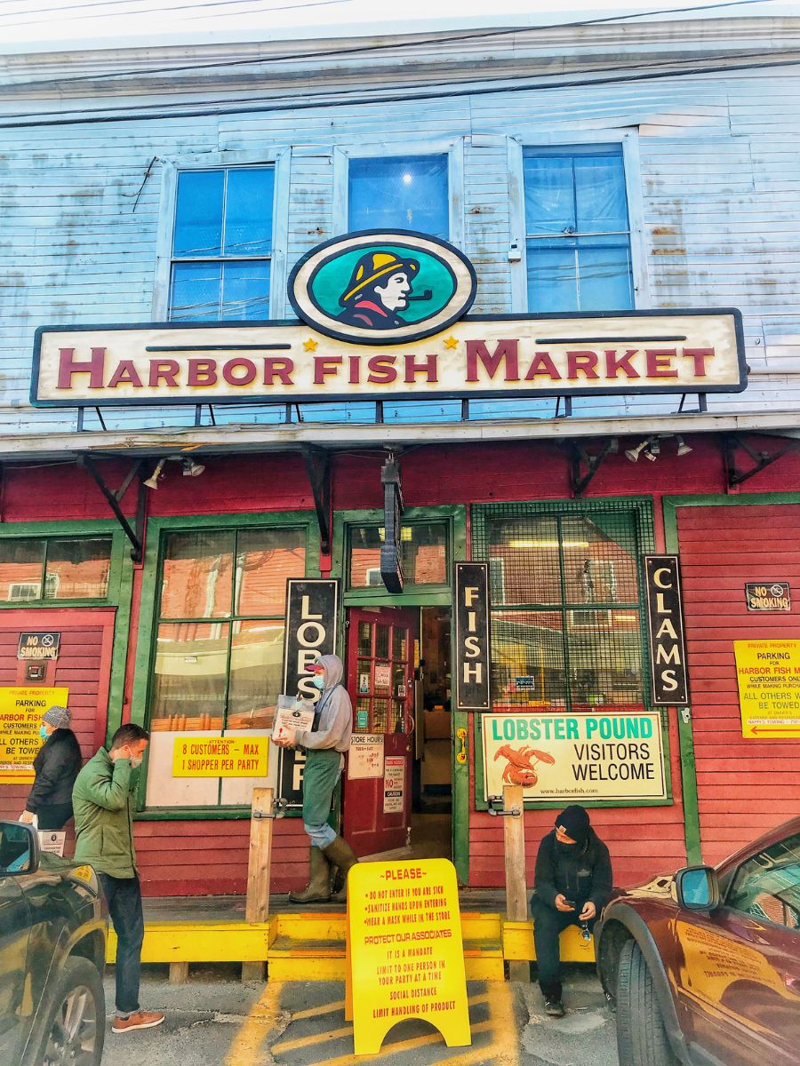 The exterior of Harbor Fish Market in Portland, Maine, with a colorful storefront and signs for lobster, fish, and clams. A worker stands at the entrance holding a box, while a customer waits nearby. Yellow signs provide guidelines for visitors, and the market has a welcoming atmosphere for those looking to buy fresh seafood.