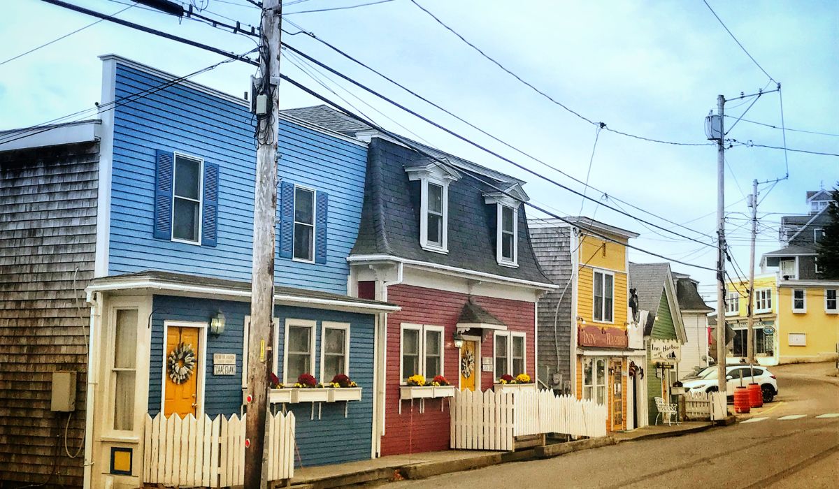 A row of colorful, charming houses in Stonington, Maine, featuring blue, red, and yellow facades with white picket fences. The quaint street is lined with overhead power lines and small businesses, capturing the cozy, small-town atmosphere.