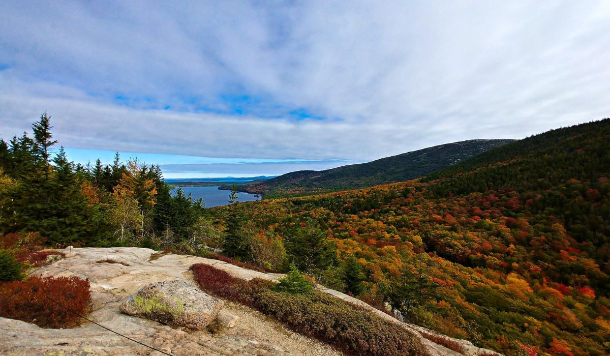 A breathtaking view of Acadia National Park in Maine, showcasing a vibrant autumn landscape with colorful foliage. The scene includes rocky terrain in the foreground, dense forests in shades of red, orange, and yellow, and a serene lake surrounded by rolling hills under a partly cloudy sky.