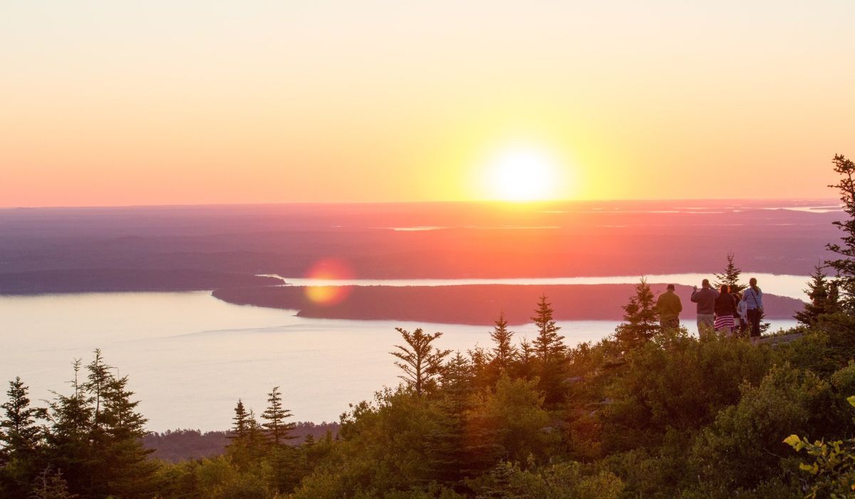 A beautiful sunrise at Cadillac Mountain in Acadia National Park, with the sun rising over the horizon and casting a warm glow over the landscape. A group of people stands among the trees, admiring the breathtaking view of the ocean and surrounding hills.