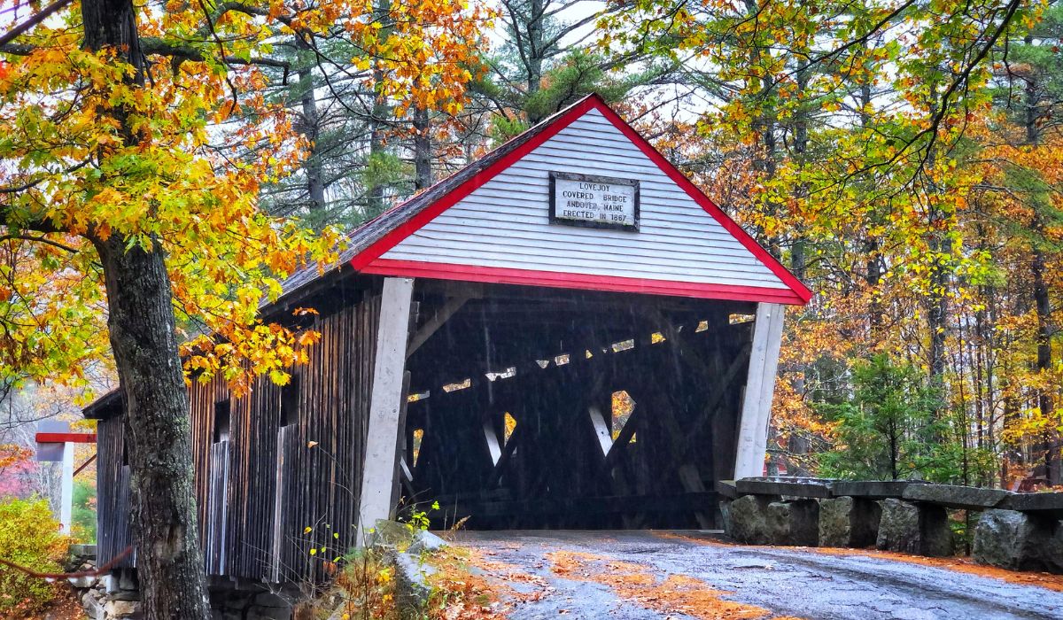 The Lovejoy Covered Bridge in Andover, Maine, surrounded by vibrant autumn foliage. The historic wooden bridge, built in 1867, is framed by colorful trees with leaves in shades of yellow, orange, and red. A narrow road leads through the quaint, rustic structure.