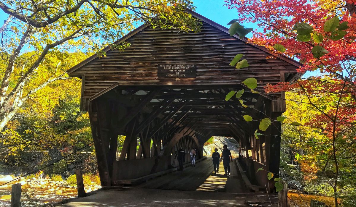 A view of the Albany Covered Bridge in the White Mountain National Forest, New Hampshire, surrounded by vibrant autumn foliage. The wooden bridge, originally constructed in 1858 and renovated in 1970, spans over a peaceful river with trees in full fall colors framing the scene. Visitors walk through the bridge, enjoying the scenic beauty and historic charm.