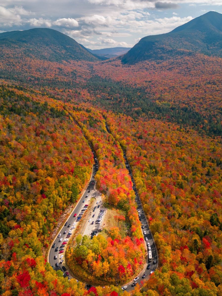 An aerial view of a scenic roadway winding through a valley in White Mountain National Forest, New Hampshire, surrounded by dense forests displaying brilliant fall colors. The mountains rise on either side, and the road is lined with parked cars as visitors enjoy the autumn foliage. The vibrant landscape is a tapestry of reds, oranges, and yellows, creating a stunning natural spectacle.