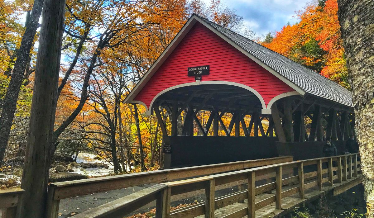 A picturesque view of the Pemigewasset River Covered Bridge in New Hampshire, surrounded by vibrant autumn foliage. The bright red bridge contrasts beautifully with the orange, yellow, and red leaves of the trees, and a wooden walkway leads to the bridge over a flowing river.