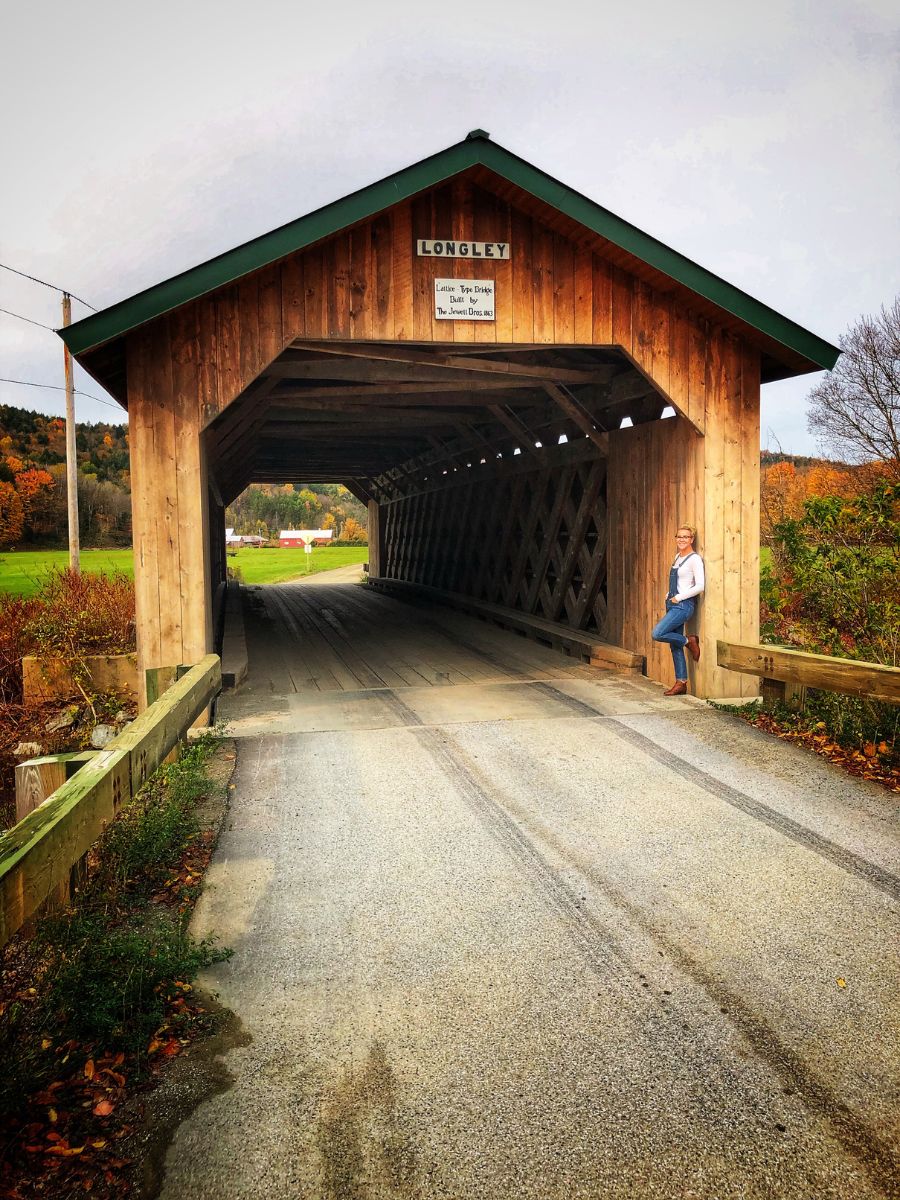 Kate stands at the entrance of the Longley Covered Bridge in Vermont, surrounded by autumn foliage. The wooden bridge stretches over a rural road with fields and trees displaying fall colors in the background.