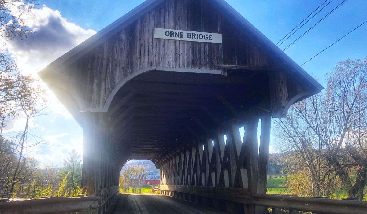 A view through the Orne Covered Bridge in Vermont, featuring its rustic wooden structure illuminated by sunlight. The bridge opens up to a pastoral landscape with green fields and distant hills under a partly cloudy sky.