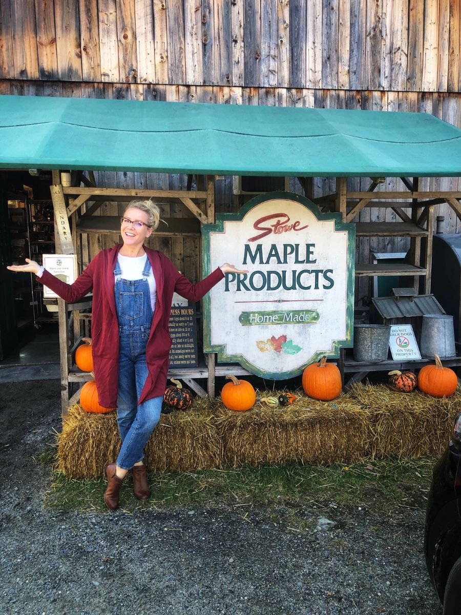 Kate stands in front of a rustic store sign for "Stowe Maple Products" in Vermont. She is wearing glasses, a white shirt, blue overalls, a red cardigan, and brown boots, with her arms spread wide in a welcoming gesture. The scene is decorated with hay bales and pumpkins, adding a festive autumnal touch.