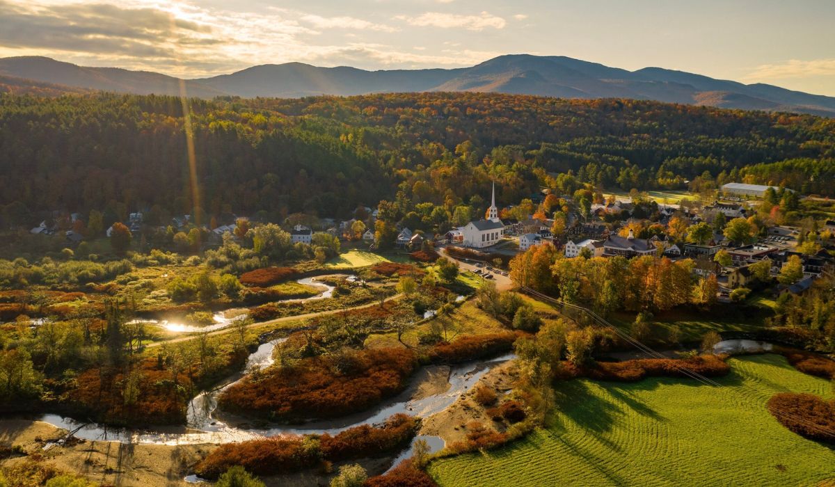 An aerial view of Stowe, Vermont, bathed in golden light during sunset. The town's iconic white church with a tall steeple is surrounded by quaint homes and colorful autumn foliage. Rolling hills and winding streams create a picturesque backdrop with mountains rising in the distance.