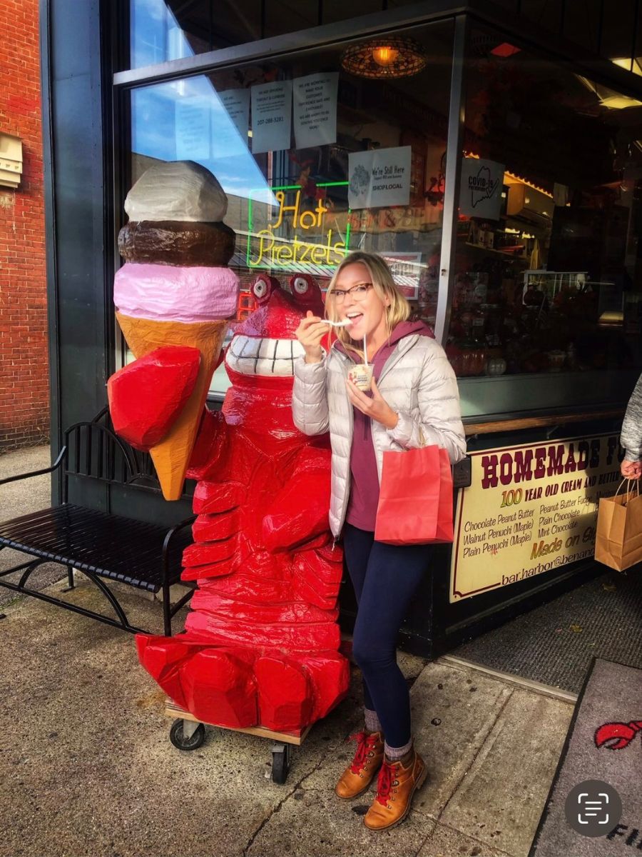 Kate stands next to a friendly red lobster statue holding an ice cream cone outside a shop. She is enjoying a cup of ice cream, wearing glasses, a light puffer jacket, and boots, holding a red shopping bag. Behind her, a neon sign reads "Hot Pretzels," and a window display advertises homemade fudge in various flavors.