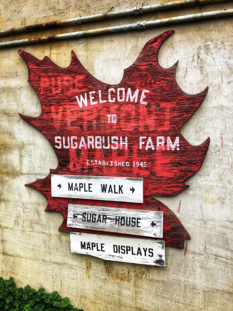 A wooden sign shaped like a maple leaf at Sugarbush Farm, Vermont, with text reading "Welcome to Sugarbush Farm Established 1945." Below, directional signs point to the "Maple Walk," "Sugar House," and "Maple Displays." The sign's rustic red paint and weathered appearance enhance the farm's charming, historical feel.