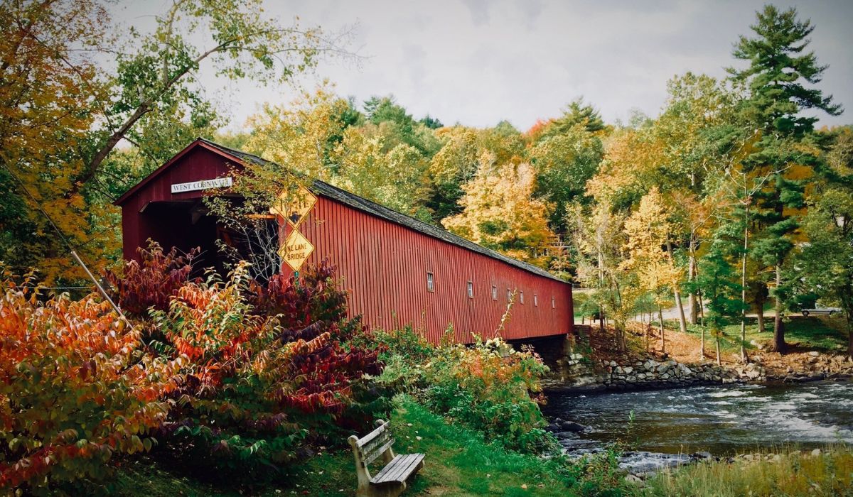 A view of the West Cornwall Covered Bridge in New England, surrounded by vibrant fall foliage with a river flowing beneath. The bridge is framed by colorful autumn leaves, with a small wooden bench in the foreground.