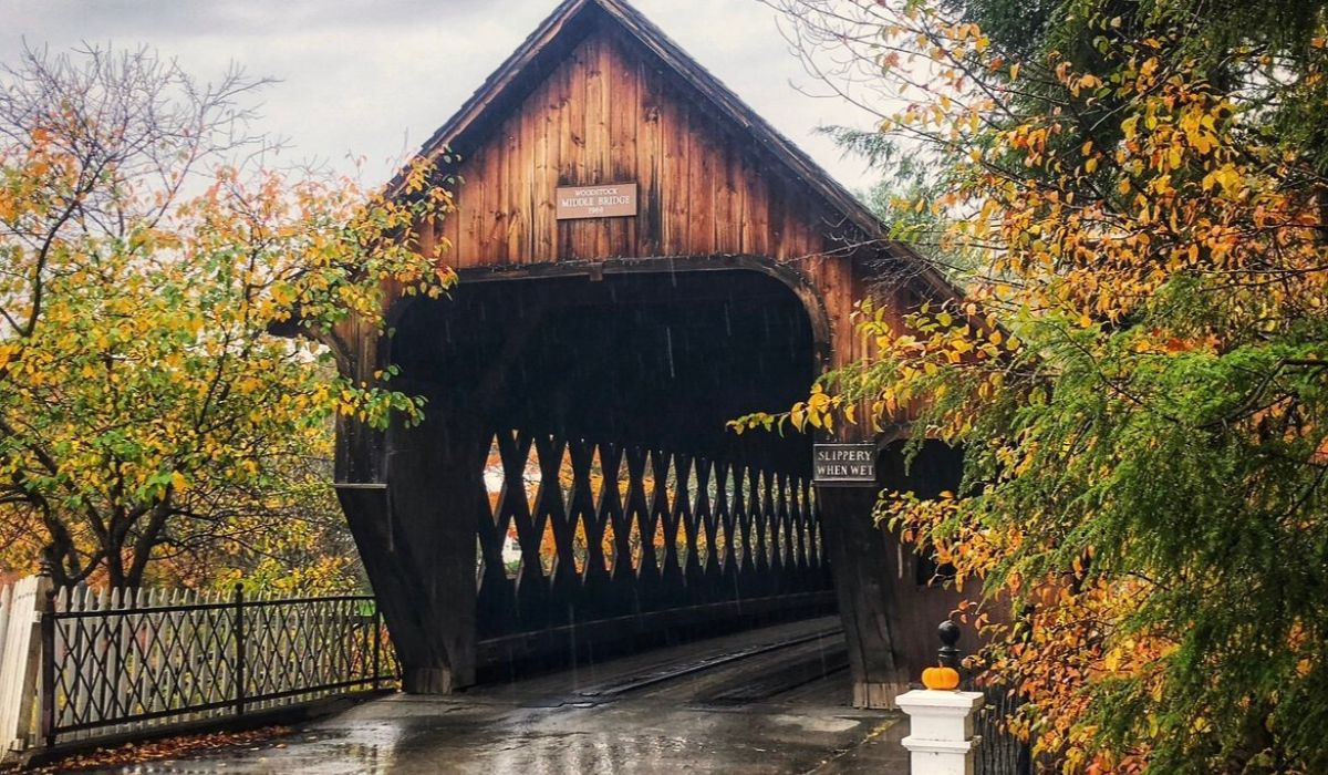 The Woodstock Middle Bridge in Vermont, surrounded by autumn foliage on a rainy day. The wooden covered bridge, adorned with a sign reading "Slippery When Wet," has a charming rustic appearance.