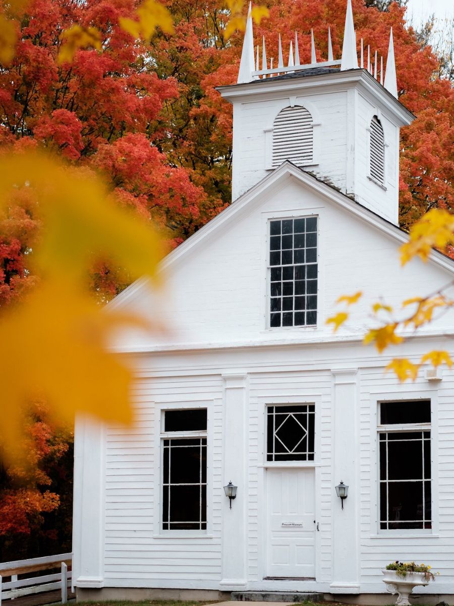 A quaint white church stands against a backdrop of vibrant red and orange autumn foliage. Yellow leaves frame the foreground, adding a touch of seasonal color to the scene. 