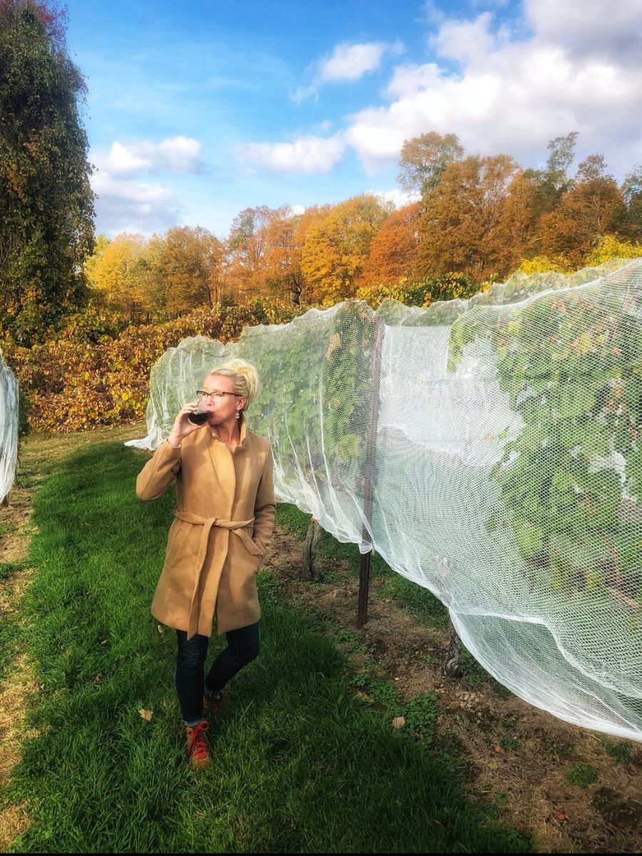 Kate stands in a vineyard during autumn, sipping wine and wearing a tan coat with a belt. The grapevines are covered with protective netting, and the background features colorful fall foliage under a partly cloudy sky.